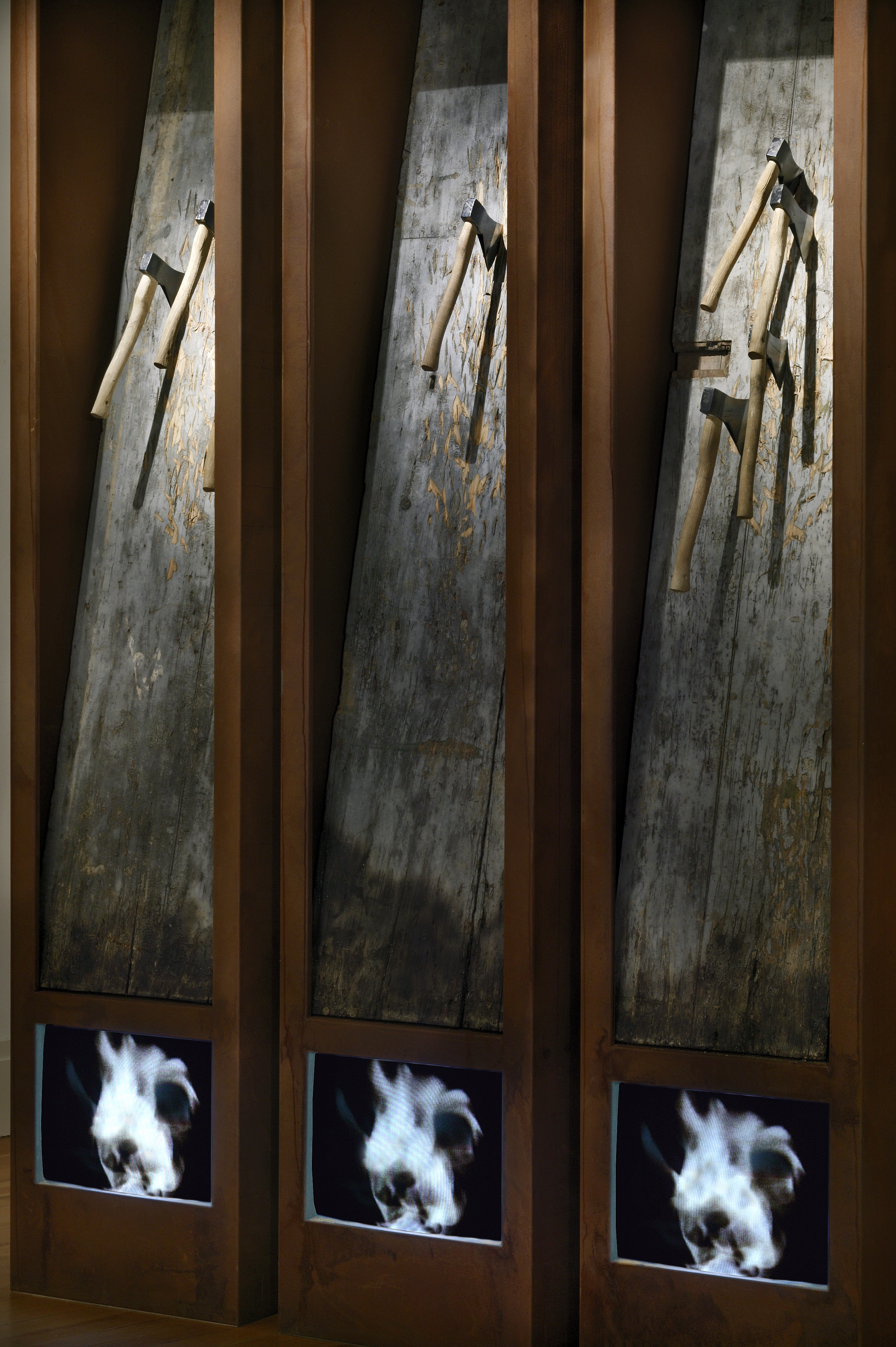 Erőszak triptichon (Trittico della violenza), installáció, 3 db rozsdásított felületű fémszekrény, 300 x 60 x kb.50 cm, a szekrényekben: faajtók, 9 db balta, 3 monitor, 2002. © Archivio PlessiTriptych of violence (Trittico della violenza), installation, 3 pieces of metal chests with corroded surface, 300 x 60 x kb. 50 cm, wooden doors, 9 axes, 3 screens built in the chests, 2002 © Archivio Plessi Trittico della violenza - I